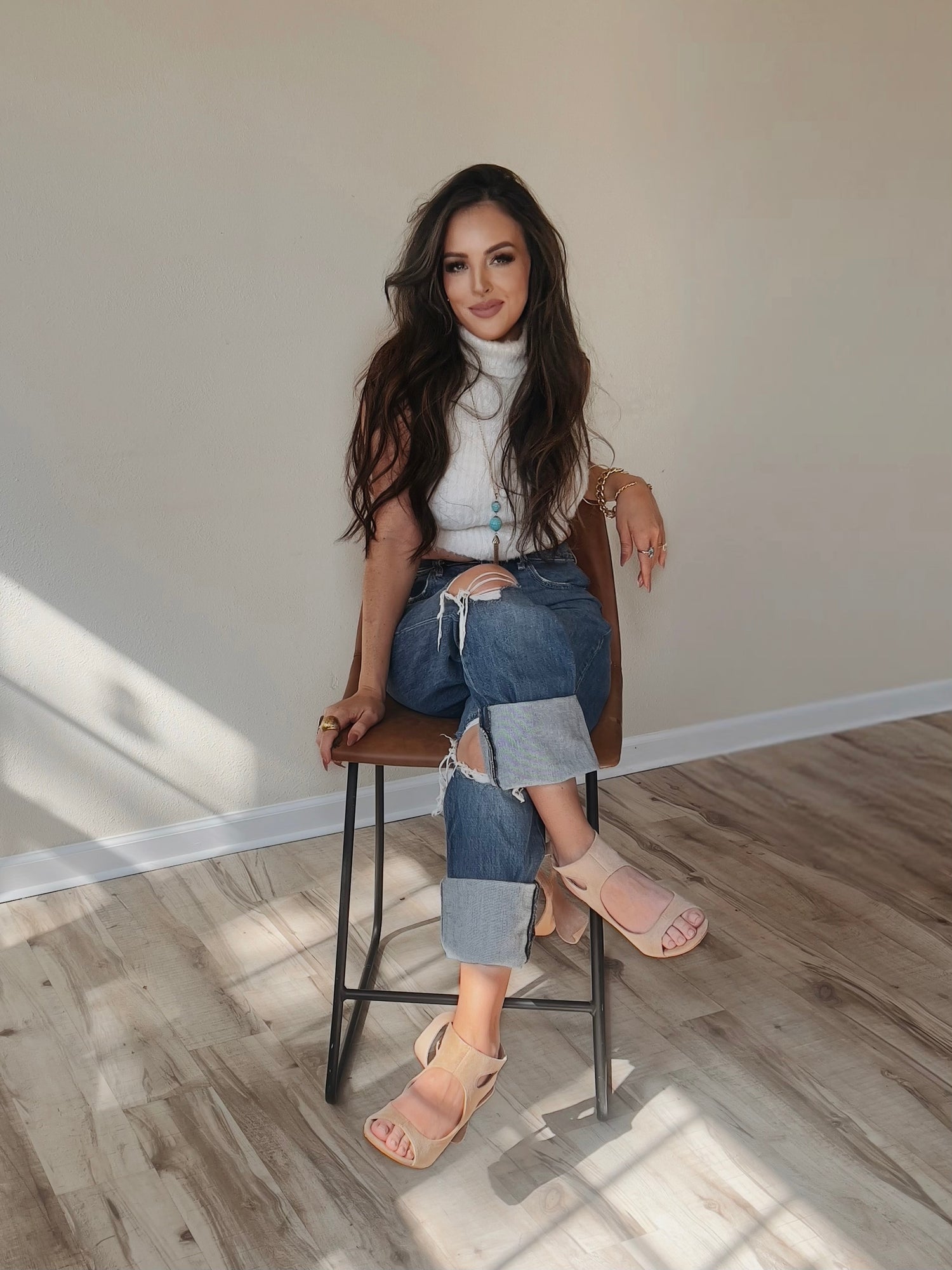 Woman sitting on a stool wearing a white top, blue jeans, and pink sandals in a room with wooden flooring.
